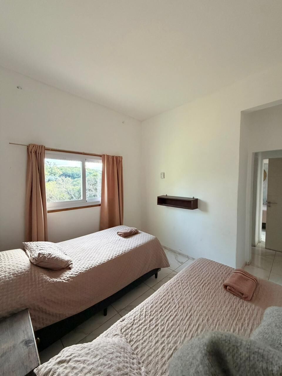 A room with two beds, a window with curtains, a wall-mounted shelf, and a door leading to another room.