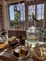 Una mesa con un pastel, un tarro de mermelada, una tetera y un vaso de agua, con vistas a la vegetación y un edificio al fondo.