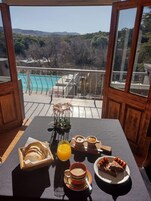 Una mesa de desayuno con pan, zumo de naranja, café y bollería, con vistas a una piscina y montañas.