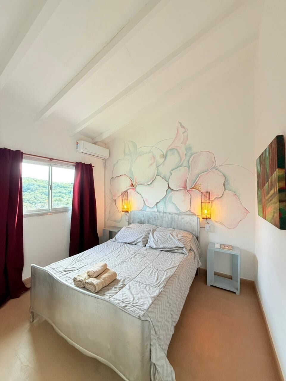 A bedroom with a bed, two bedside tables, a wall-mounted light fixture, and a window with curtains.