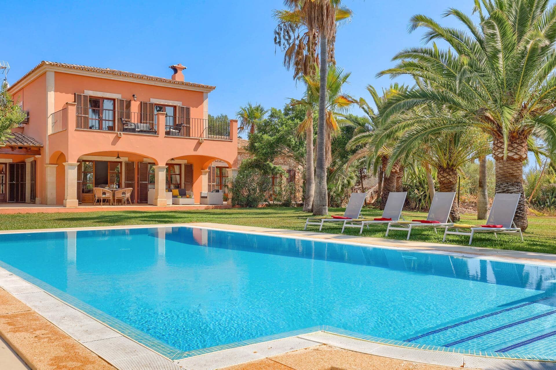 A two-story house with a swimming pool, lounge chairs, and palm trees.