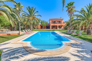 A swimming pool with lounge chairs, palm trees, and a building with a red facade. - Villa Marino by Villa Plus (Santañy)
