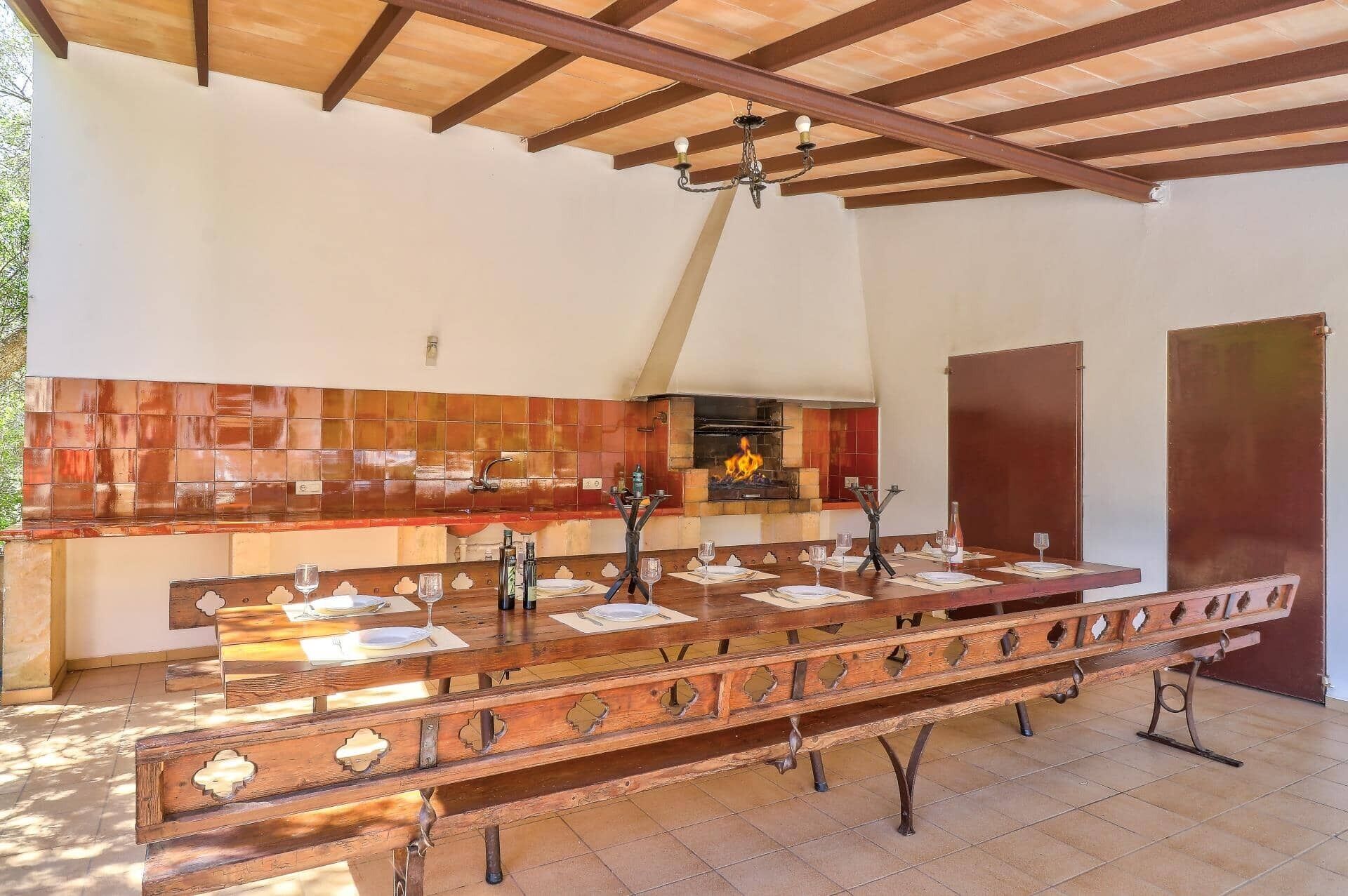 Une longue table à manger en bois, avec des chaises, un coin cuisine doté d’un mur recouvert de carreaux, et une cheminée.