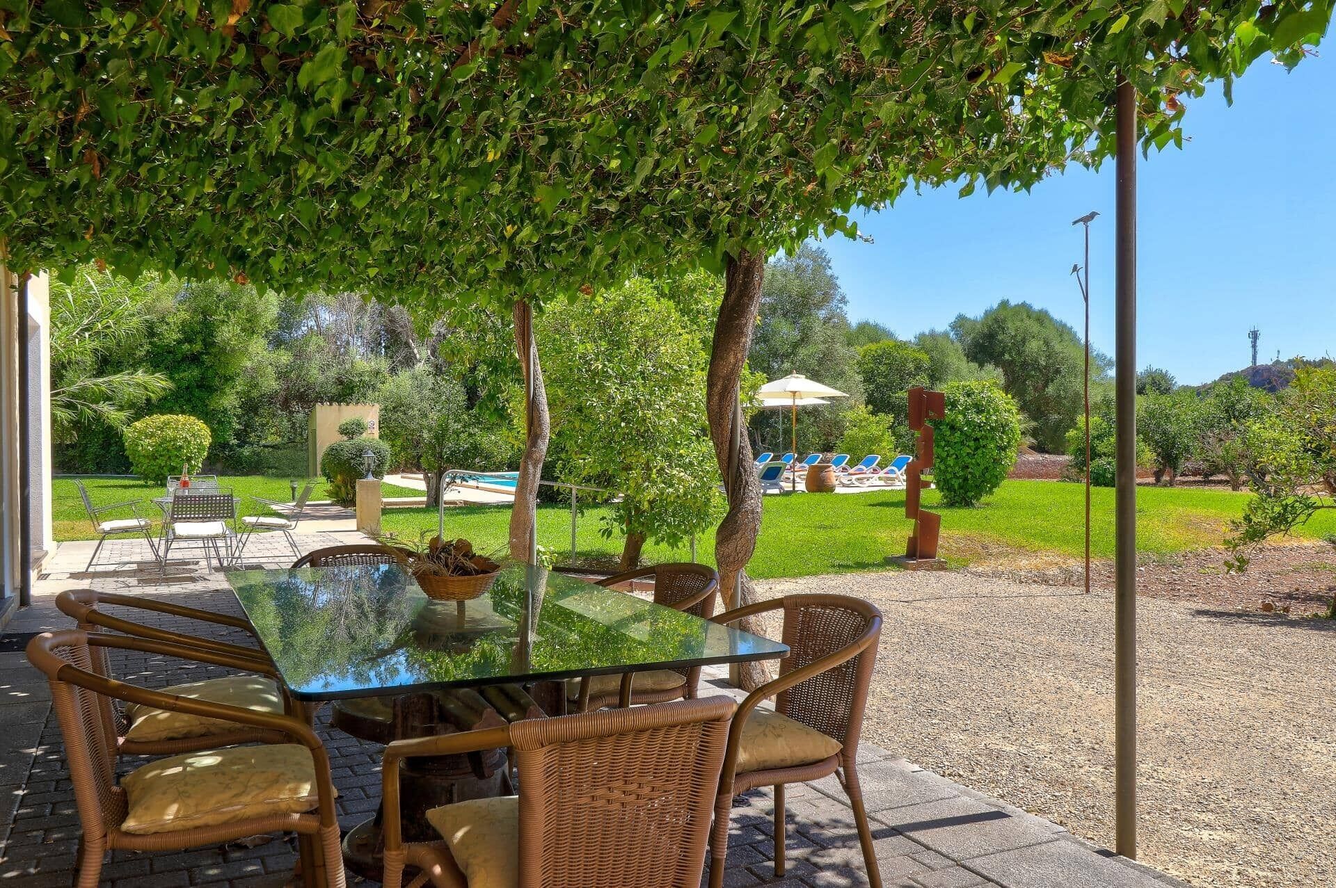 Un espace de restauration en plein air avec une table en verre et des chaises en osier, entouré de verdure et avec une piscine en arrière-plan.