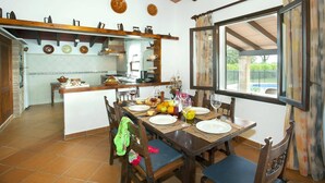 A dining area with a wooden table set for four, surrounded by chairs. A kitchen is visible in the background with a hanging pot rack above the counter.