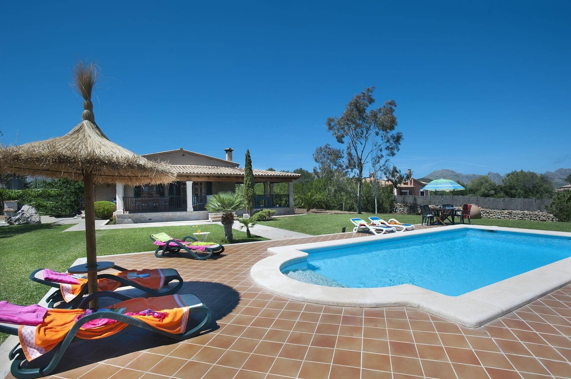 A swimming pool with lounge chairs and a thatched umbrella, surrounded by a well-maintained lawn and a house with a tiled roof.