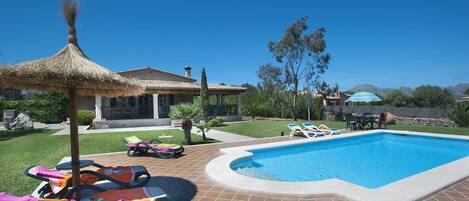 A swimming pool with lounge chairs and a thatched umbrella, surrounded by a well-maintained lawn and a house with a tiled roof.