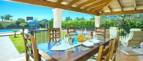 Un patio al aire libre con mesa de madera dispuesta para comer, rodeado de sillas de mimbre y vistas a una piscina y exuberante vegetación.