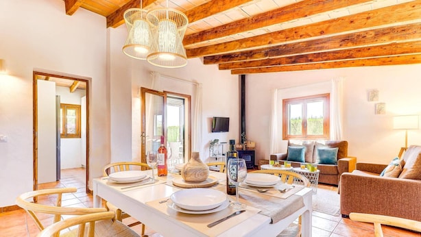 A dining area with a white table set for four, surrounded by wicker chairs. A wooden ceiling with exposed beams and a hanging light fixture is visible. - Villa Olivo by Villa Plus (Mallorca)