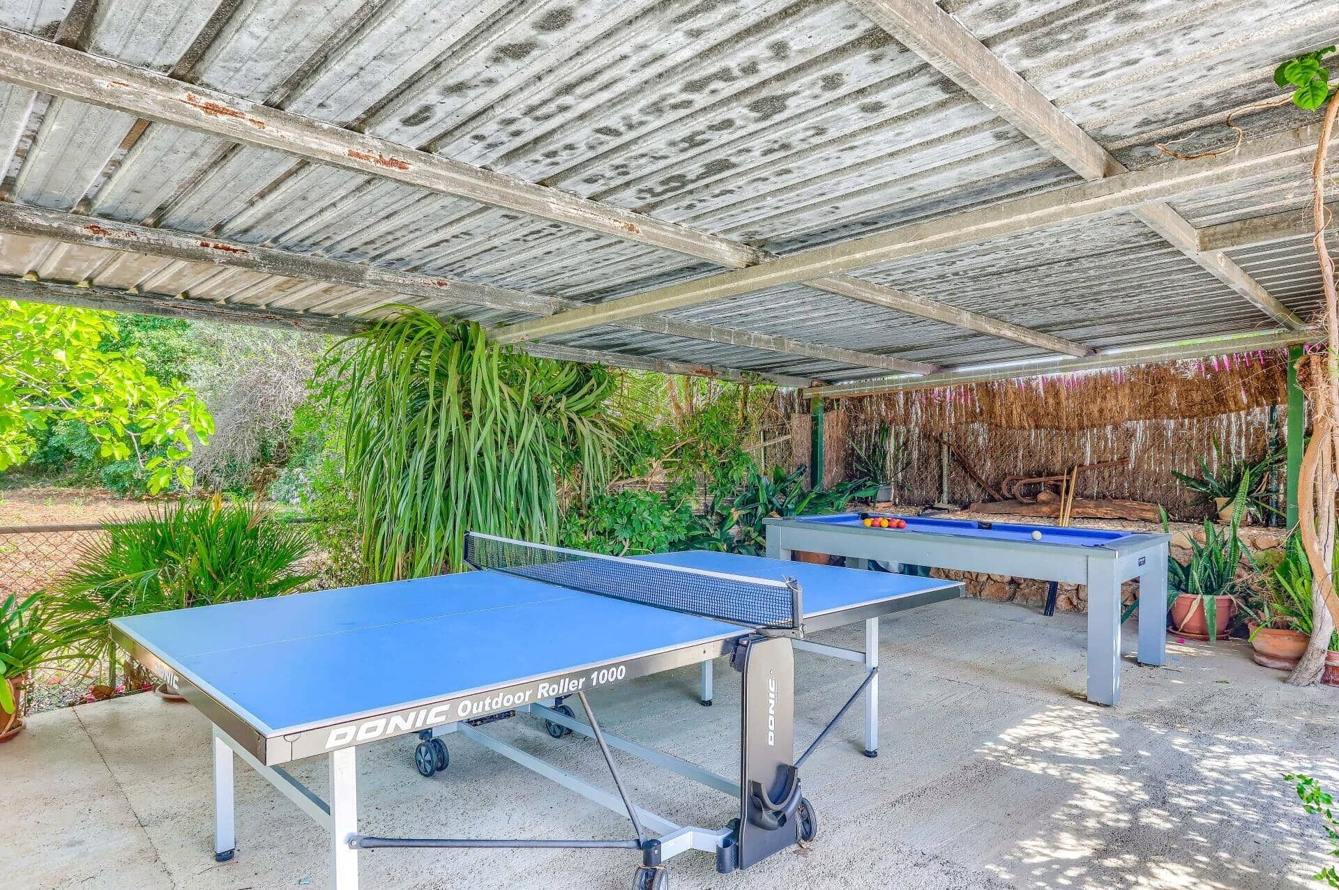 Two outdoor ping pong tables under a corrugated metal roof with greenery around.