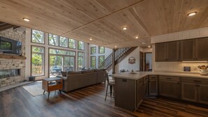 Amplia sala de estar con chimenea de piedra, ventanas grandes, techo de madera y una cocina con piso de madera oscura.