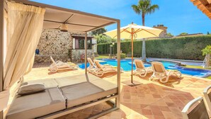 A poolside area with lounge chairs, a covered seating area, and a stone wall.