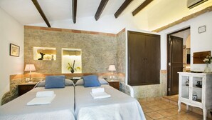 Una cama doble con dos almohadas, una mesita de noche con una lámpara, una pared de piedra, una puerta de madera y un mueble pequeño con un jarrón.