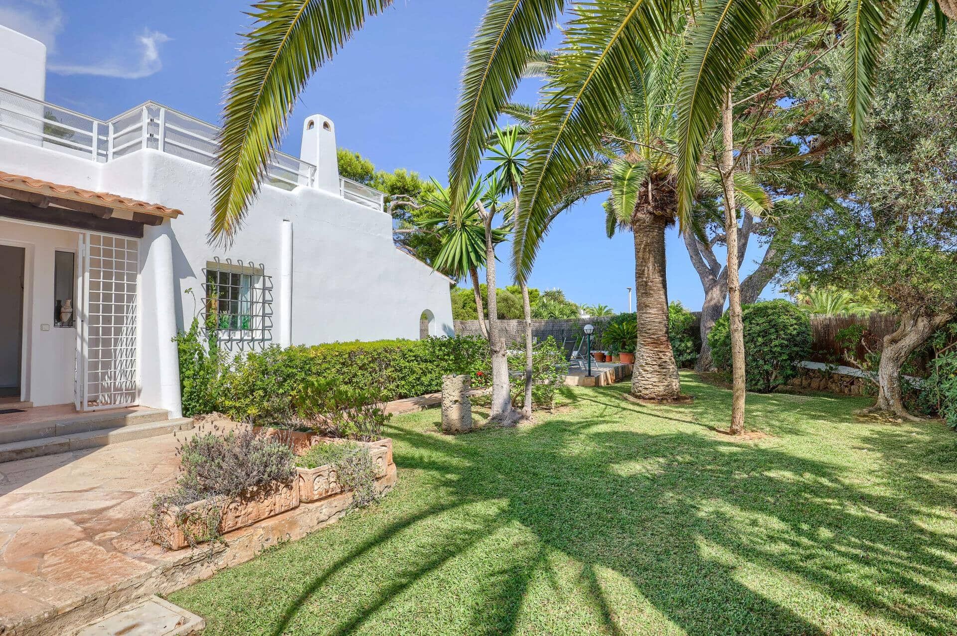 Un bâtiment blanc doté d’un balcon, entouré d’un jardin bien entretenu où l’on trouve des palmiers et des arbustes.