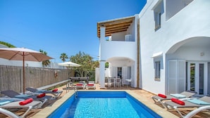 A swimming pool with lounge chairs and umbrellas, a wooden deck, and a white building with a balcony.