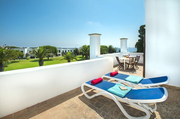 Ein Balkon mit zwei Liegestühlen, einem Tisch mit Stühlen und Blick auf das Meer und Gebäude im Hintergrund.