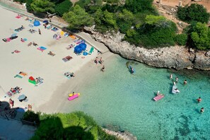 Luchtbeeld van een strand met helder turquoise water, een zandstrand en mensen die zonnebaden en zwemmen.