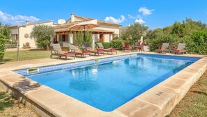 A swimming pool with lounge chairs and a covered seating area, surrounded by greenery and a building with a tiled roof. - Villa Garriguer by Villa Plus (Mallorca)