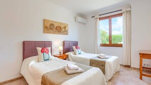 A hotel room with two beds, a window with curtains, an air conditioning unit, and a painting on the wall. - Villa Garriguer by Villa Plus (Mallorca)