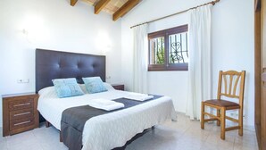 A bedroom with a bed, wooden nightstands, a chair, a window with curtains, and a view of greenery outside. - Villa Francisca by Villa Plus (Mallorca)