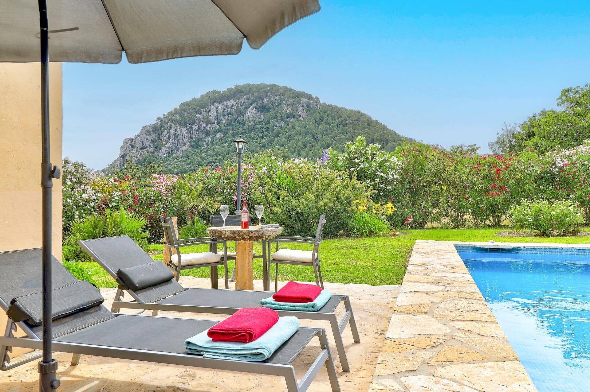 A poolside area with lounge chairs, a table with a lamp, and a view of a mountainous landscape.