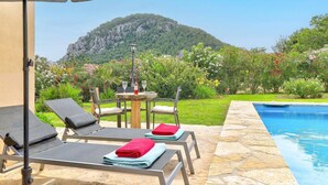 A poolside area with lounge chairs, a table with a lamp, and a view of a mountainous landscape.