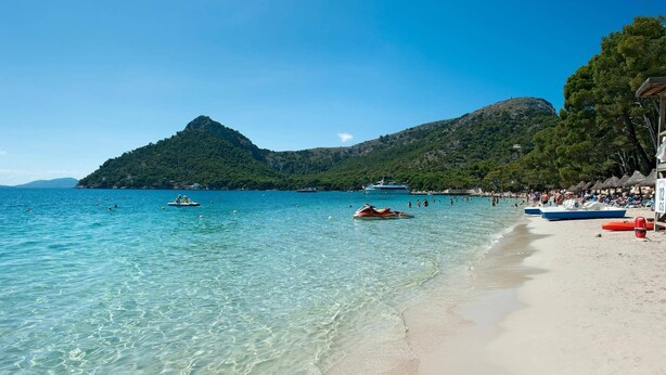 A beach with clear turquoise water, a sandy shore, and a mountainous backdrop.