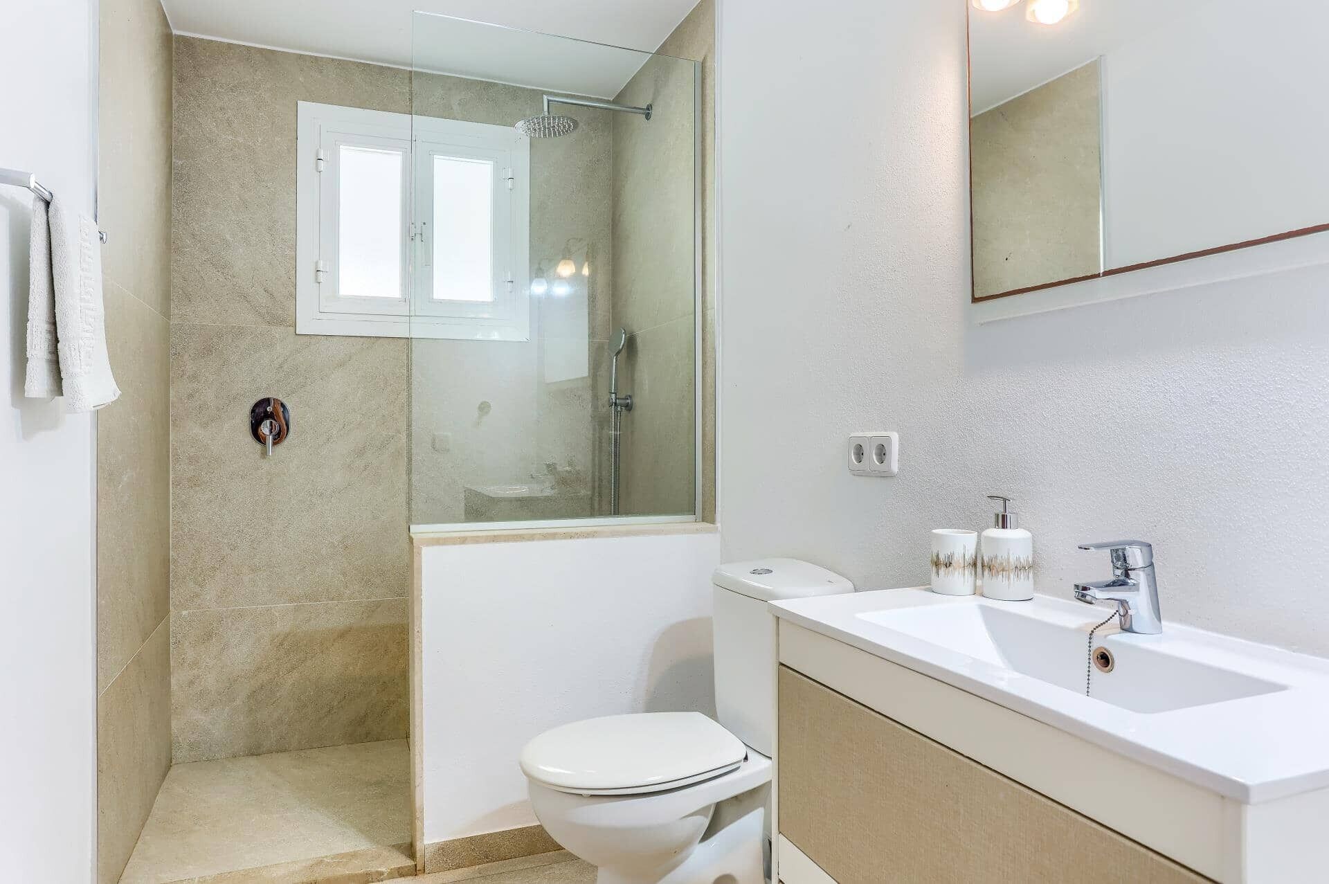 Un baño moderno con ducha envuelta en vidrio, inodoro blanco y tocador con espejo y dos dispensadores de jabón.