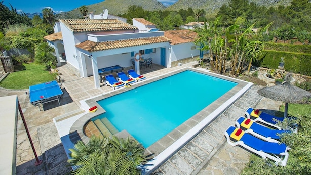 Un espace aménagé au bord d’une piscine à l’eau d’un bleu limpide, avec des chaises longues et un bâtiment doté d’un toit recouvert de tuiles.