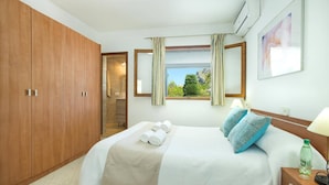Une chambre d’hôtel avec un grand lit, une armoire, une salle de bain visible par une porte ouverte, une fenêtre avec des rideaux et un climatiseur fixé au mur.
