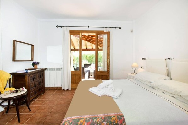 A bedroom with a bed, a bedside table with a lamp, a wooden dresser, a chair, a window with curtains, and a view of the outdoors.