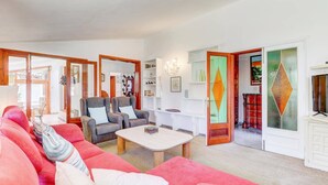 Un vaste salon avec un canapé rouge, des fauteuils gris et une table basse. On y trouve également un coin repas avec une table et des chaises rouges, et une cuisine visible à travers une porte ouverte.