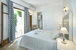 A bedroom with a bed, a nightstand, a lamp, a chair, and a bathroom visible through an open door.