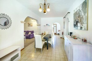 A spacious living area with a dining table, a purple sofa, and a white armchair. There is a chandelier hanging from the ceiling and a large abstract painting on the wall.