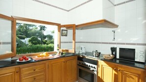 A kitchen with wooden cabinets, a stove, a microwave, and a window overlooking a garden. - Villa Malen by Villa Plus (Cala d'Or)