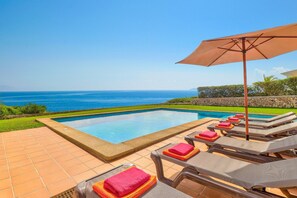 A poolside area with lounge chairs and an umbrella, overlooking a clear blue ocean. - Villa S'Algar by Villa Plus (Felanitx)