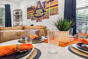 A dining table set with plates, glasses, and orange napkins, with a plant and framed pictures on the wall. - NEWLY RENOVATED, 3 blocks from AU, Game Day House “Caddy’s Shack” (Auburn)