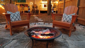 Två trästolar i Adirondack-stil med rutiga kuddar, en eldstad med brinnande ved och en stensatt gång som leder till en träveranda med ljusslingor.