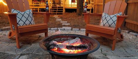 Due poltrone in legno di stile Adirondack con cuscini a quadretti, un braciere con legna ardente e un vialetto in pietra che conduce a un terrazza in legno con luci a stringa.