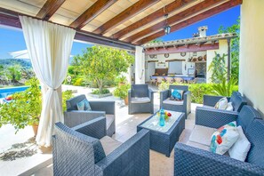 Un espacio de estar techado al aire libre con muebles de mimbre, una piscina y una cocina al fondo.