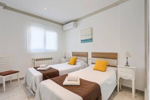 A hotel room with two beds, a window with curtains, a radiator, a bedside table with a lamp, and a painting on the wall.