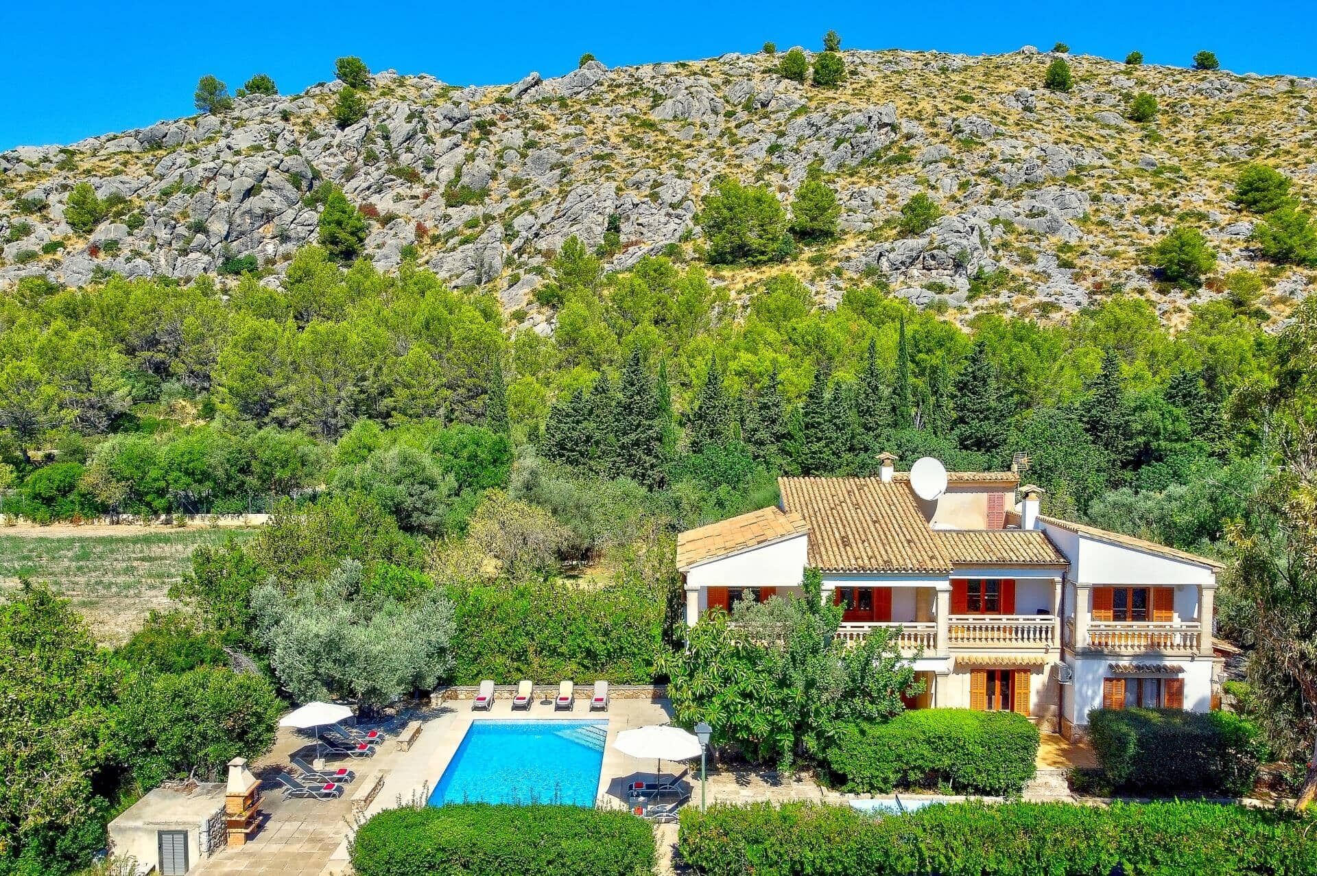 A two-story house with a swimming pool, surrounded by lush greenery and a rocky hillside.