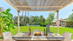 Un'area pranzo all'aperto con una pergola, una piscina e un giardino curato sullo sfondo.