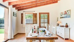 Zona pranzo con tavolo in legno, sedie e un armadietto bianco. Si vedono finestre con tende, un quadro appeso al muro e uno scorcio di verde all'esterno.