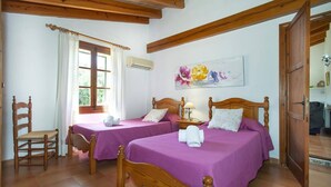A room with two wooden beds, a chair, a window with curtains, an air conditioning unit, and a floral painting on the wall. - Villa Vidrier II by Villa Plus (El Vilà)