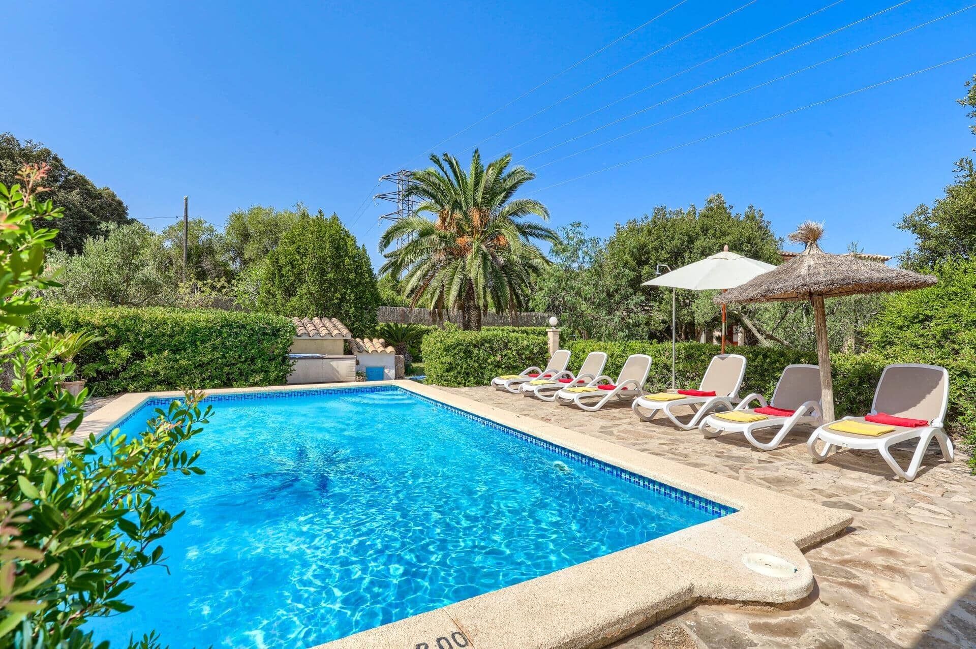A clear blue swimming pool with lounge chairs and umbrellas, surrounded by greenery and palm trees.