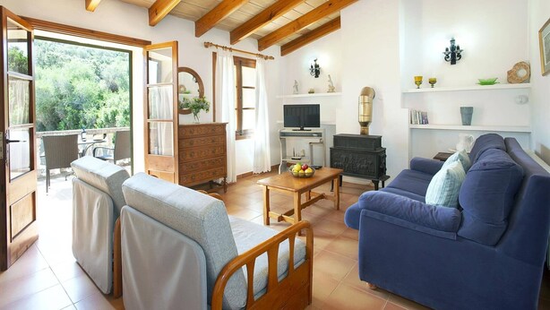 A cozy living room with a sofa, a wooden coffee table, and a television. There is a fireplace and a window overlooking greenery.