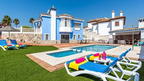 A poolside area with lounge chairs, a swimming pool, and a two-story building with blue and white exterior walls. - Villa Tia Ana by Villa Plus (Fuengirola)