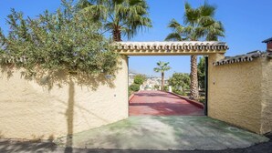A gated entrance with a red pathway leading to a building, surrounded by palm trees and a clear sky. - Villa Tia Ana by Villa Plus (Fuengirola)