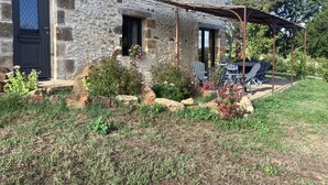Ein Steinhaus mit einer dunklen Tür, einem kleinen Fenster mit Blumen und einem überdachten Sitzbereich mit Stühlen und einem Tisch.